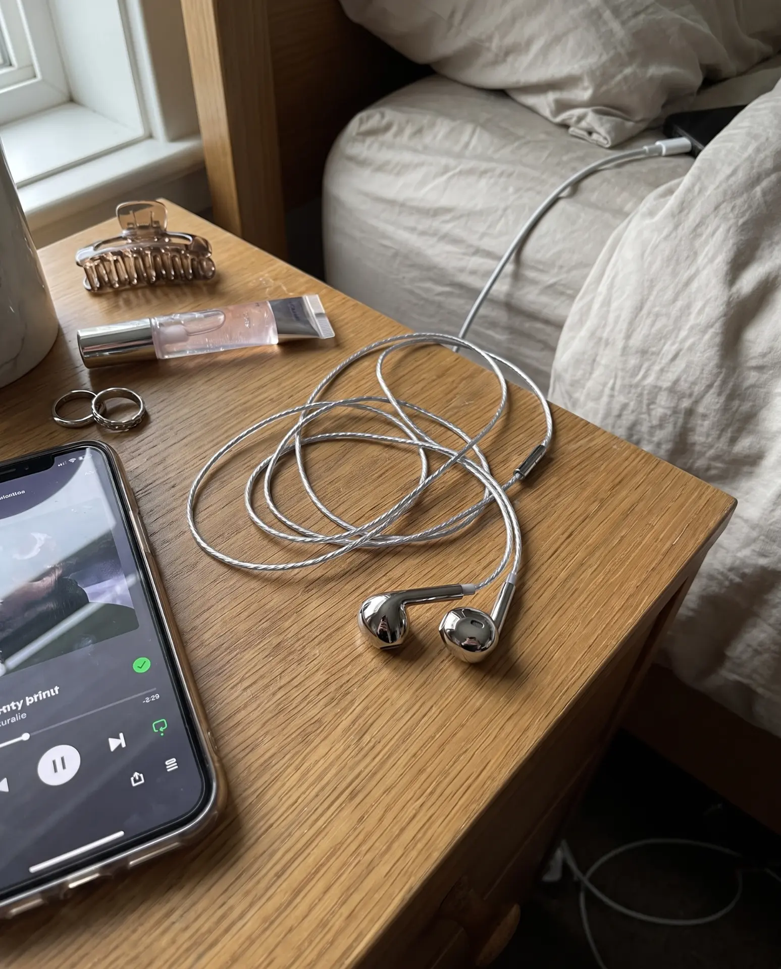 Silver Auralie earbuds coiled on a wooden nightstand beside rings, lip gloss and a phone playing music