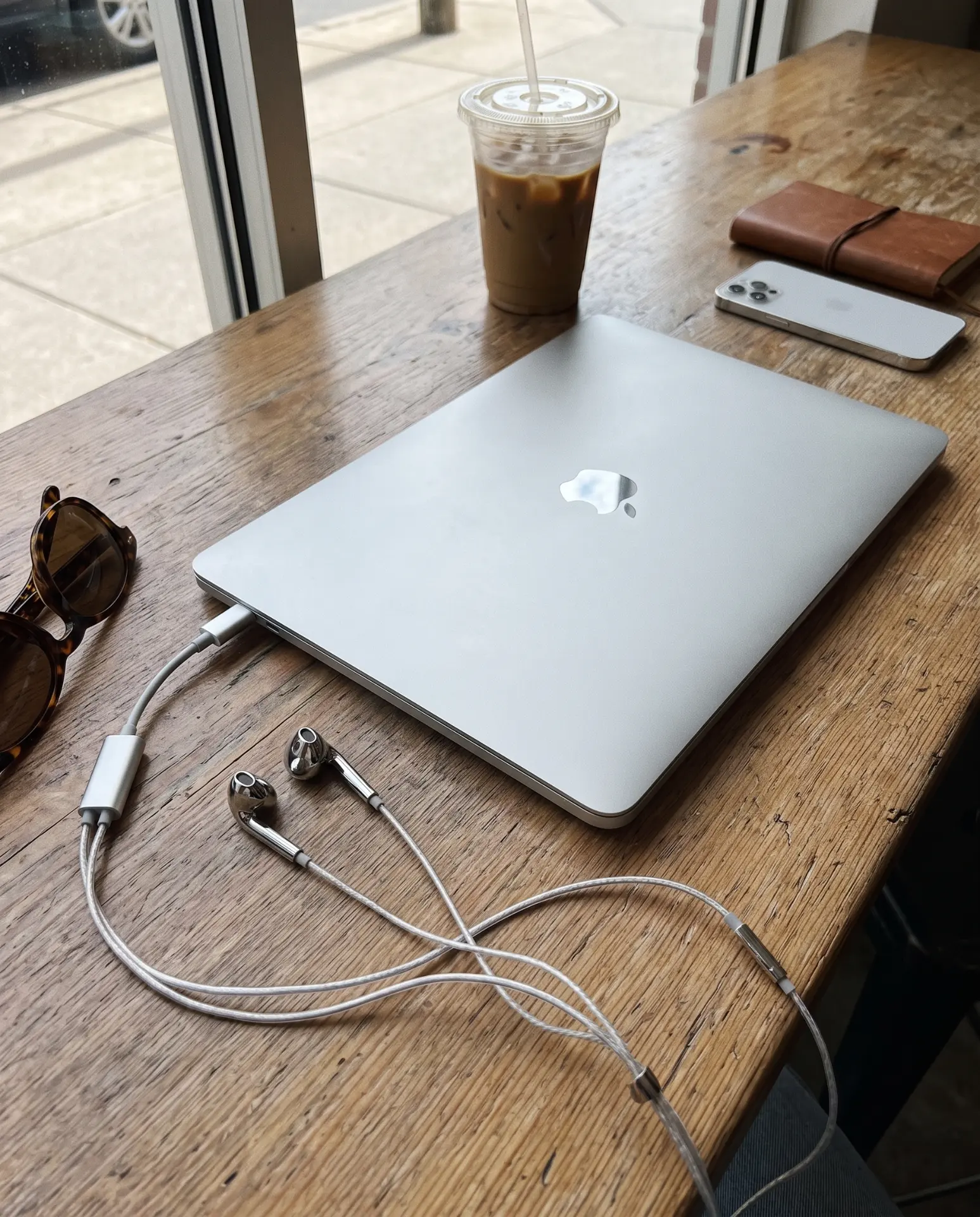Silver Auralie earbuds plugged into a laptop at a café table with an iced coffee and sunglasses