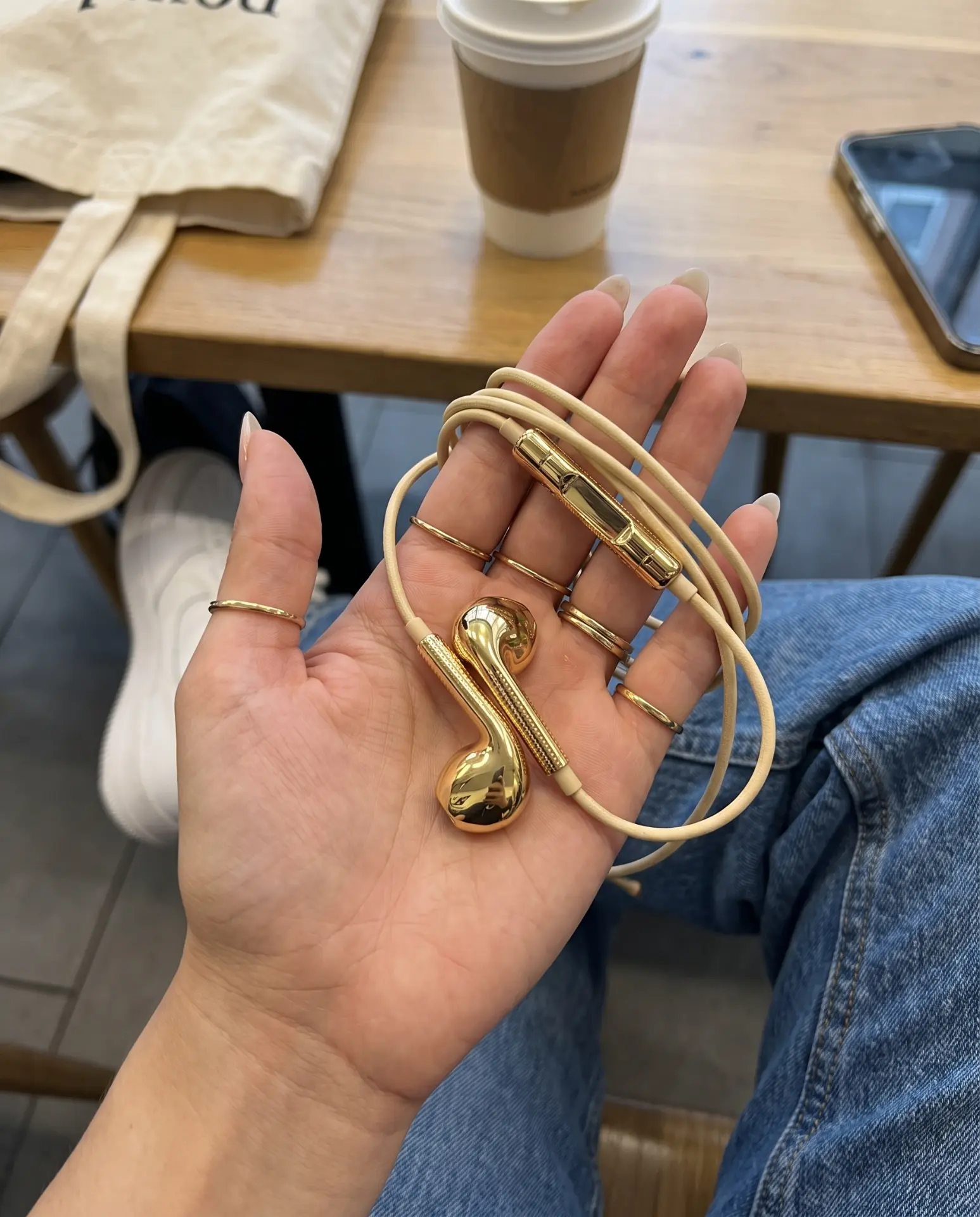 Gold Auralie earbuds held in a hand with rings, café table and tote bag behind