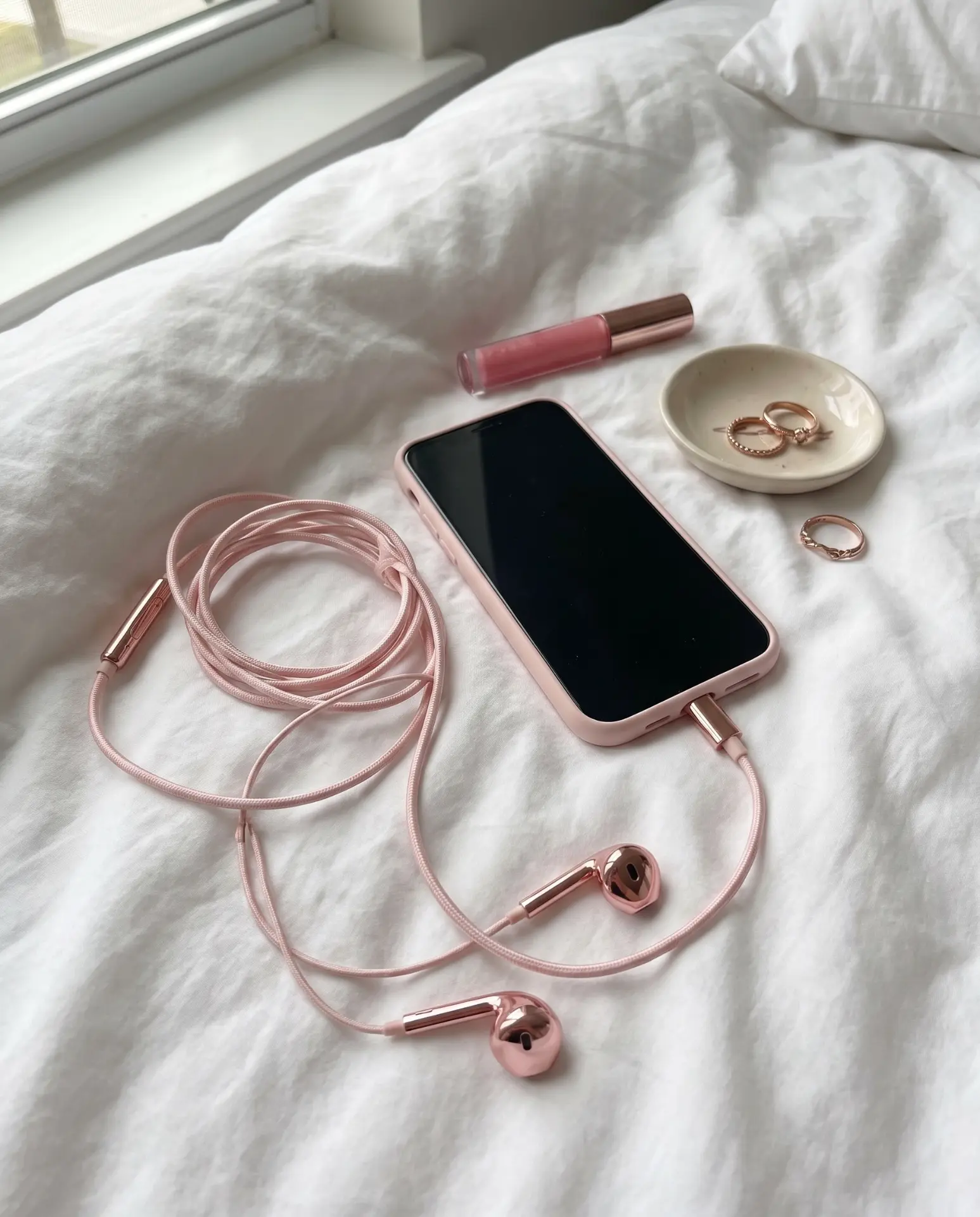 Rose gold Auralie earbuds and a phone laid flat on a bed with lip gloss and rings